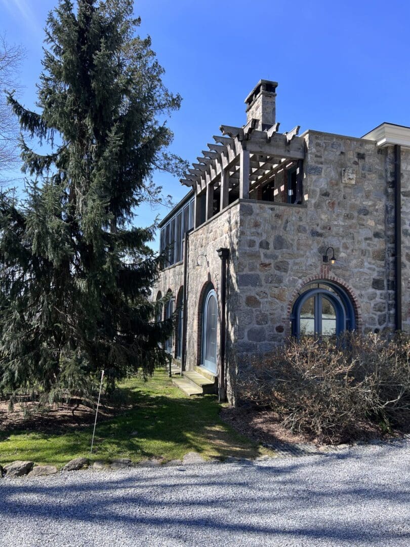 Stone building with arched windows beside a tall pine tree on a sunny day.