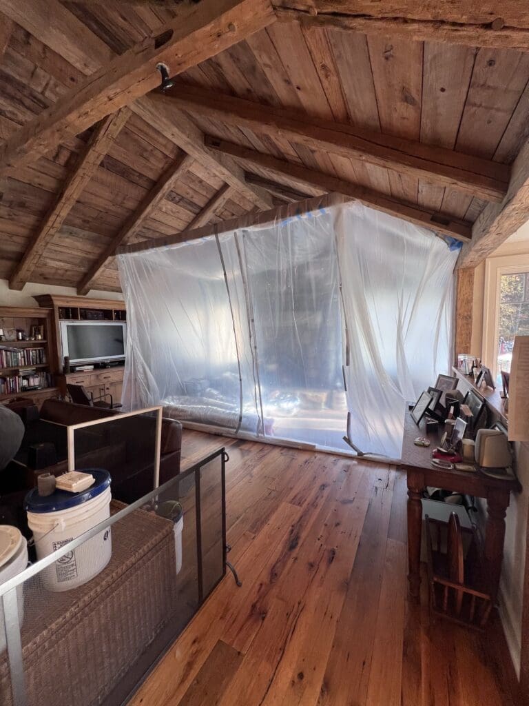 Plastic sheeting encloses an area under a wooden ceiling in a rustic room.
