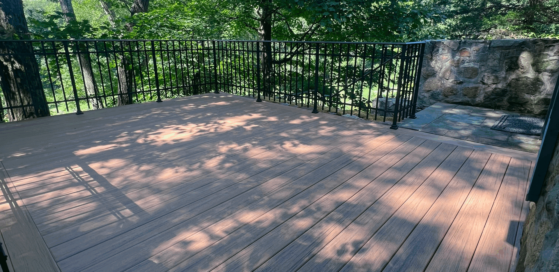 Sunlit wooden deck with black metal railing surrounded by greenery.