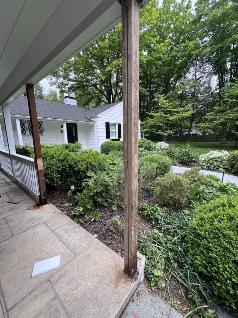 A porch with a missing wooden post overlooks a garden and a white house.