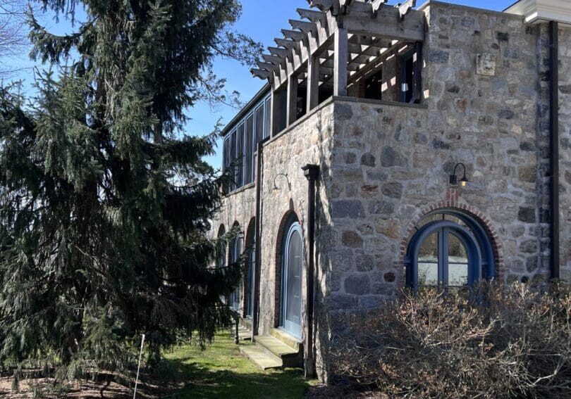 Stone building with arched windows beside a tall pine tree on a sunny day.